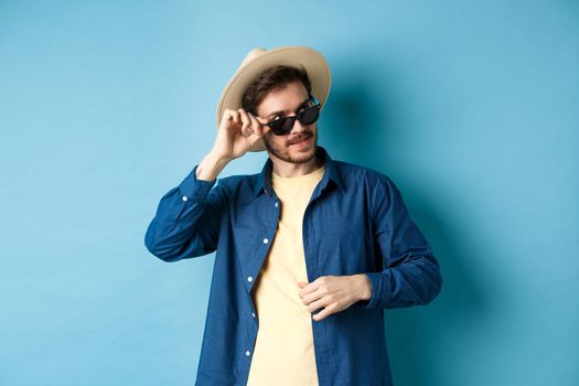 Handsome tourist in sunglasses and straw hat looking aside, smiling pleased, exploring sighsteeing on vacation, blue background.