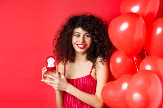 Pretty caucasian woman getting engaged on Valentines day. Girl receive marriage proposal on lovers holiday, showing golden ring in small box, standing near hearts balloon on red background.