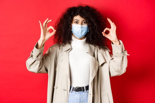 Covid-19, social distancing and quarantine concept. Fashionable woman with curly hair, wearing medical mask and trench coat, showing okay gestures, red background.