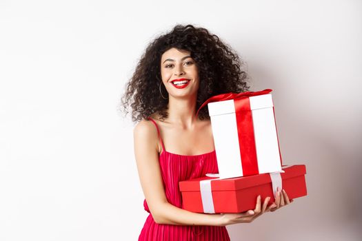 Beautiful birthday girl with curly hair, holding bday gifts and smiling happy, celebrating, standing against white background.