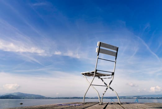 View of a lonely and empty chair with a landscape of a calm lake with blue sky and light clouds. With copy space in the upper left side. Empty concept, absence.