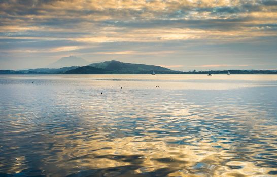 View of Lake Zugersee in the Swiss town of Zug at sunset with beautiful golden tones in the sky and reflected in the water and clouds.