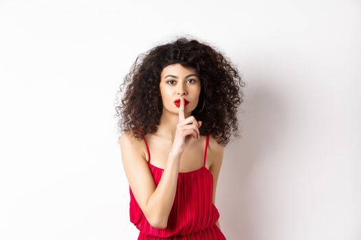 Beautiful woman with curly hair, wearing red dress, hushing at camera, telling secret, shushing at you, standing over white background.