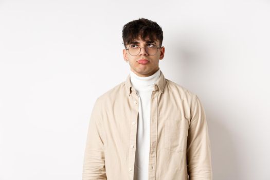 Image of sad handsome guy feeling disappointed, pouting lips and looking at empty space distressed, standing on white background in stylish clothes and eyewear.