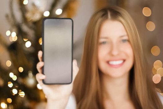 Christmas phone call and holiday greeting concept. Happy smiling woman showing screen of mobile smartphone, xmas decoration on background.