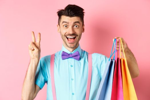 Cheerful guy showing peace sign and shopping bags, smiling happy at camera, standing over pink background.