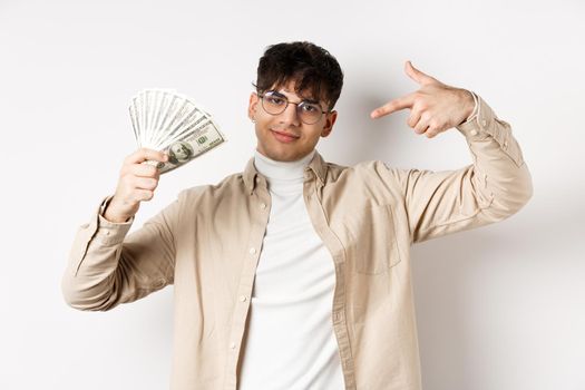 Cool handsome guy show-off his income, pointing at dollar bills and smiling boastful, making money, standing on white background.