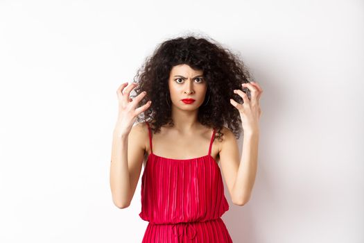 Angry italian woman with curly hair and red dress, boiling from anger, frowning and clenching hands outraged, standing on white background.