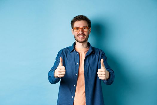 Happy young man in glasses showing thumbs up, recommending optician store, standing on blue background.