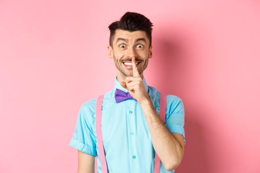 Cheerful young man making surprise, shushing at camera with happy smile, asking to keep voice down, be quiet, standing on pink background.