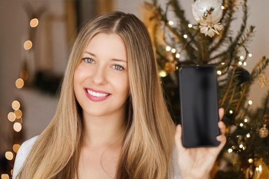 Christmas phone call and holiday greeting concept. Happy smiling woman showing screen of mobile smartphone, xmas decoration on background.