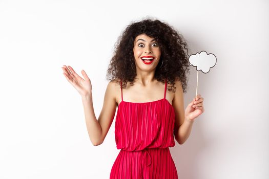 Happy young woman in red dress, holding comment cloud and looking surprised, standing over white background. Copy space