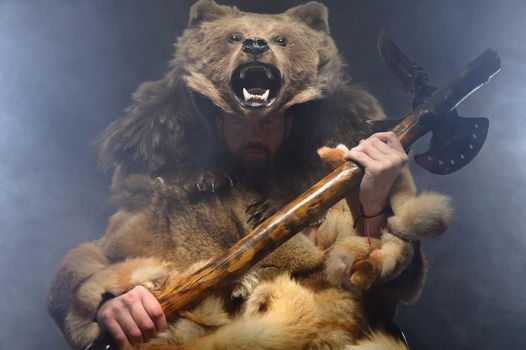 Portrait of a man Bearded Scandinavian shaman with axe on hands and a bear skin on his head. Caucasian man dressed in animal skins pensively looks at camera