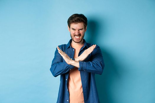 Angry young man frowning and clenching teeth outraged, showing cross gesture to stop or forbid something, standing on blue background.