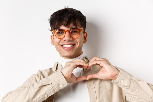 Close-up of young natural guy in glasses smiling, showing heart love you gesture, standing on white background.