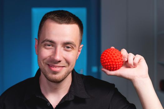 Portrait of a young Caucasian physiotherapist masseur on a blue background with a red massage ball in his hands. Smiling and looking at the camera. Self-massage and self-treatment concept.