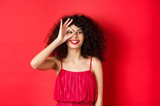 Beautiful woman look through okay sign with excitement, checking out promo offer, standing in redd against red background.
