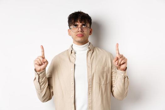 Image of annoyed or offended young man looking, pointing fingers up bothered, frowning disappointed, standing on white background.