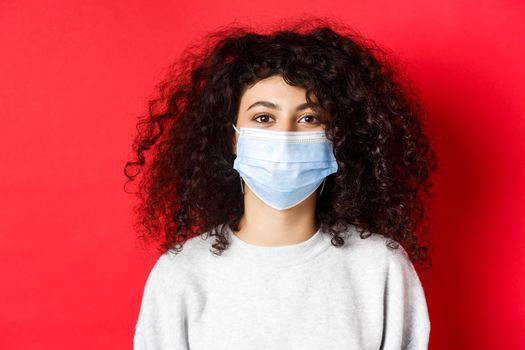 Covid-19 and pandemic concept. Close-up of modern young woman with curly hair, wearing medical mask from coronavirus, smiling at camera, red background.