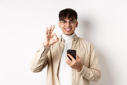 Technology and online shopping concept. Portrait of handsome modern guy in glasses showing OK gesture using smartphone, recommending app or shop, white background.