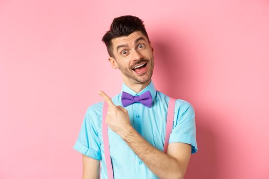 Funny male model in bow-tie, tilt head and pointing finger left, inviting to look, check it out gesture, showing advertisement on pink background.