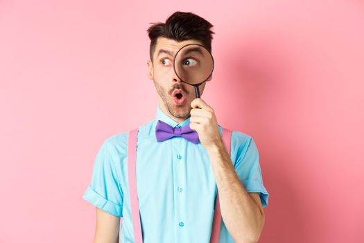 Surprised man saying wow and look aside through magnifying glass, checking out promo offer, standing over pink background. Copy space