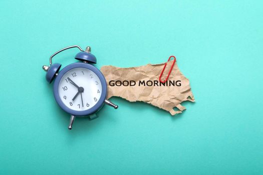 Conceptual photo of blue alarm clock with business card and text good morning on paper background.