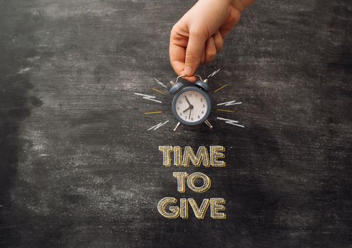 Chalk text time to give on black school board. At the top of the board, a female hand holds an alarm clock. Donation concept.