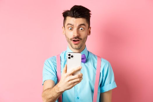 Impressed man reading mobile screen with amazed face, seeing online promo offer, standing over pink background. Copy space