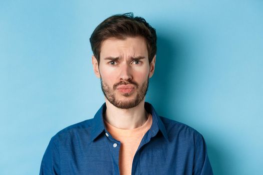 Close up of sad and offended young man sulking at someone, frowning upset, standing on blue background.