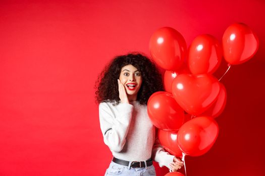 Valentines day. Surprised curly-haired girl holding heart balloons and looking amazed at camera, receive surprise gift on date from lover, standing on red background.