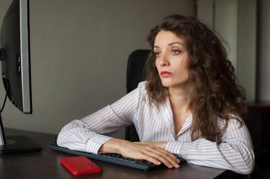 Tired young woman with curly hair and white shirt is working at the office using her laptop, routine work, freelance, burnout syndrome.