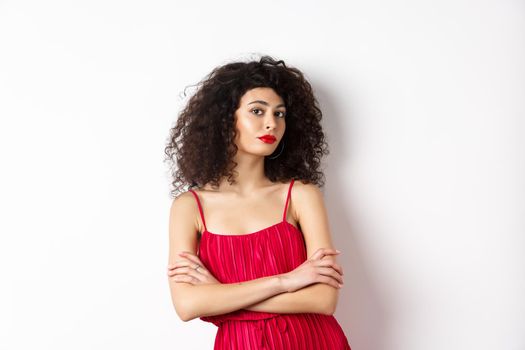 Sassy woman with curly hairstyle, wearing red dress and makeup, cross arms on chest, standing over white background.