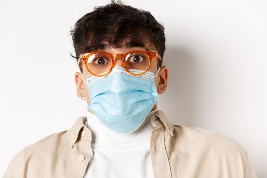 Coronavirus pandemic and healthy people concept. Surprised guy in glasses and face mask raising eyebrows, stare at camera in awe, standing on white background.