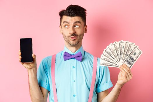 E-commerce and shopping concept. Funny guy look curious at empty smartphone screen, showing money, buying online, standing on pink background.