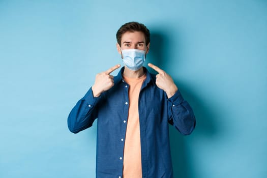 Coronavirus and pandemic concept. Portrait of happy and healthy man pointing fingers at his medical mask, smiling at camera, standing on blue background.