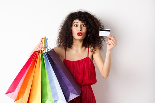 Excited woman in red dress, holding shopping bags and showing plastic credit card, shop with discounts, white background.