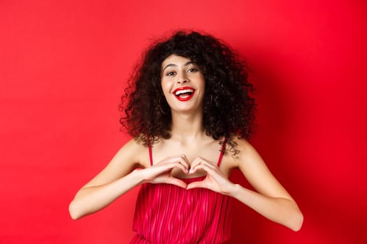 Beautiful woman with curly hair, red dress, showing heart symbol and smiling happy, studio background.