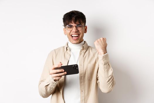 Happy guy winning on smartphone, holding mobile phone and raising hand up, shouting yes with joy, standing on white background.