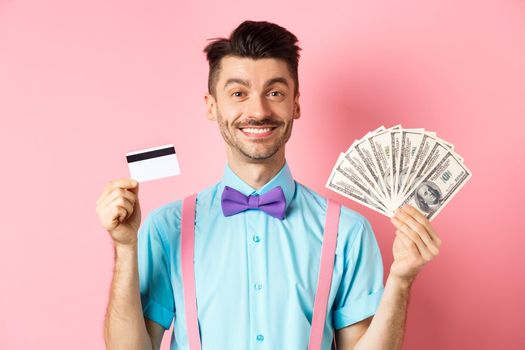 Cheerful man showing his money and plastic credit card, smiling happy at camera, standing over pink background.