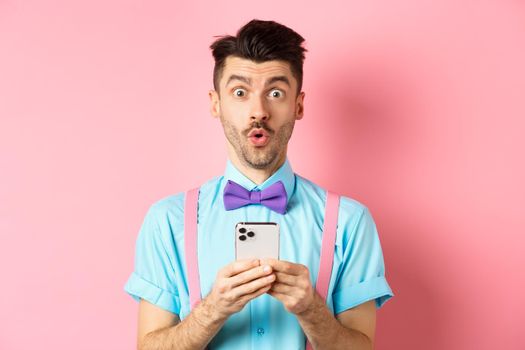 Online shopping. Intrigued guy in bow-tie, checking out internet promo offer on mobile phone, say wow at camera, holding smartphone, standing on pink background.