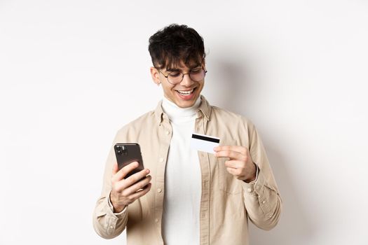 Happy young guy in glasses making online purchase, looking at plastic credit card and holding smartphone, standing on white background.