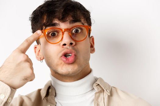 Close-up of cute and funny guy in glasses pointing at eyewear frames, recommending optic store, standing on white background.