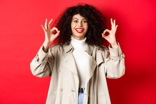 Stylish caucasian woman in trench coat showing okay gesture and smiling satisfied, recommending company, standing on red background.