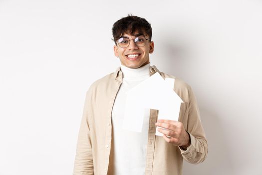 Real estate and mortgage concept. Cheerful young guy renting apartment, showing paper house cutout and smiling happy, buying property, white background.