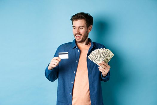 Handsome young man showing cash and looking at plastic credit card, prefer contactless payment, standing on blue background.