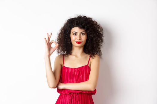 Beautiful assertive woman with curly hairstyle and makeup, wearing red dress, showing okay sign and smiling in approval, say yes, standing on white background.