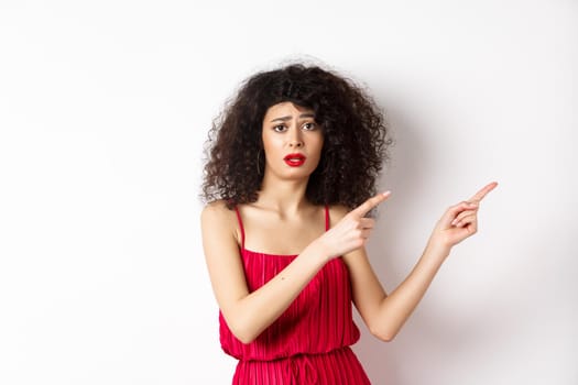 Confused and sad woman with curly hair, wearing evening dress and makeup, pointing fingers right and frowning upset, standing on white background.