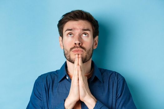 Close up shot of hopeful guy pleading God, holding hands in begging gesture and looking up in sky, standing on blue background supplicating.