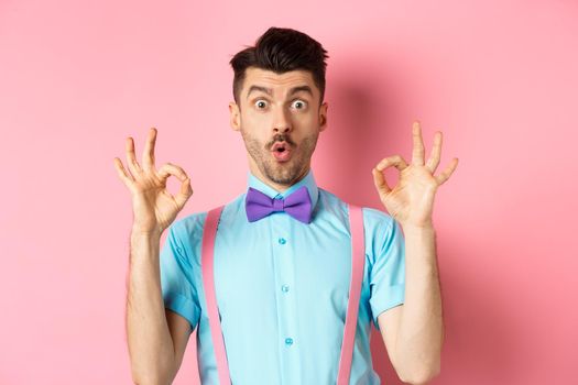 Impressed caucasian guy in bow tie, checking out cool offer, showing okay gesture and saying wow, praising good product, standing on pink background.